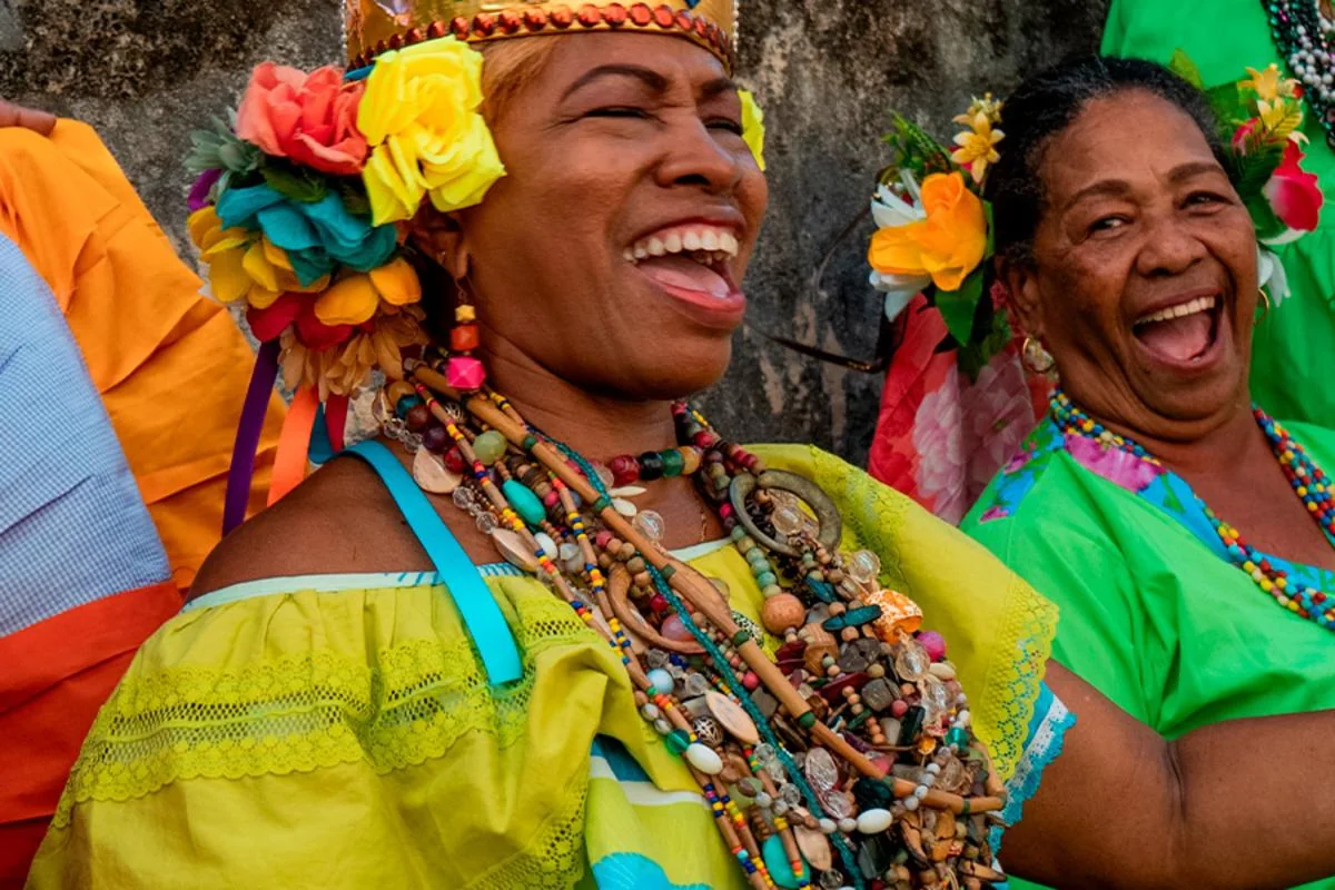 Dos mujeres riendo con flores en el pelo y collares de cuentas, vestidas para una celebración congo
