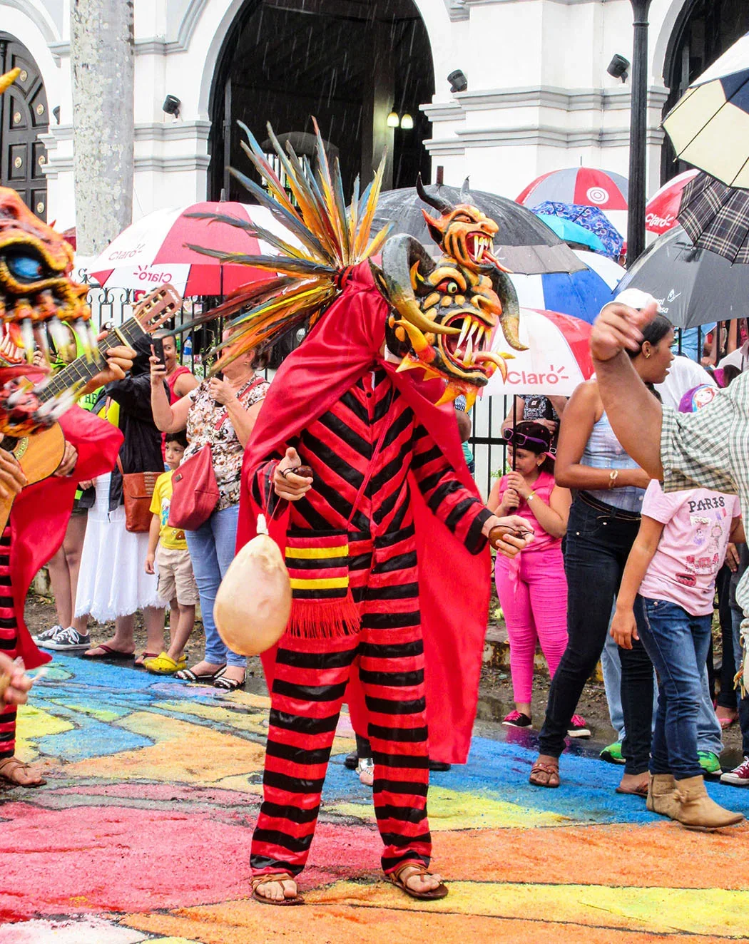 Diablo con máscara y traje rojo y negro bailando bajo la lluvia frente a una iglesia, entre espectadores con paraguas